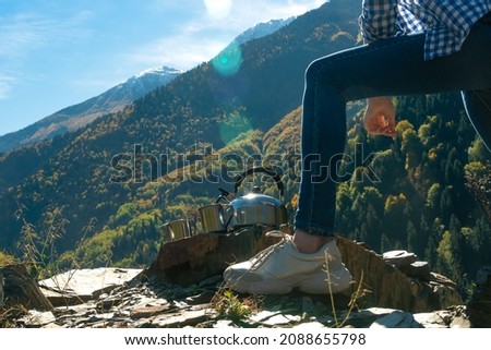 Close-up of a young woman's leg near a teapot and cups against a mountain landscape on a sunny autumn day. Concept of travel, outdoor activities, camping