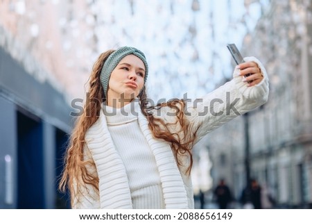 Cool posing for selfies in the city center. A young bright cute girl in a white knitted suit and a blue headband takes a selfie. Duck lip pose. Fashion photography concept.