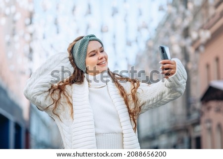 A young stylish stunning girl with long curly hair dressed in a white warm knitted suit takes a selfie in the city. A raised hand and a bright smile. The concept of a high-quality selfie.