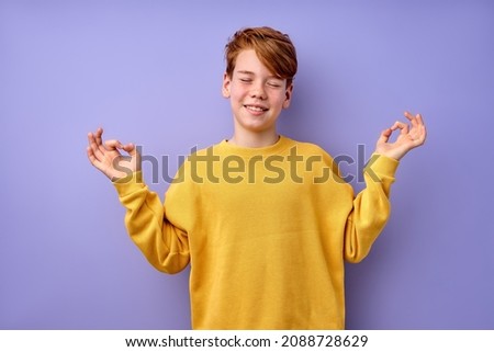 Portrait of young teenage boy keep calm, meditating, isolated on purple studio background. Kid in yellow casual shirt standing with eyes closed engaged in yoga, focused on thoughts, smiling