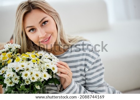 Female posing with bouquet of flowers at home. Housewife received present for holiday. Allergy free. Good-looking pretty blonde lady is relaxed, enjoying the smell, look at camera. in bright room