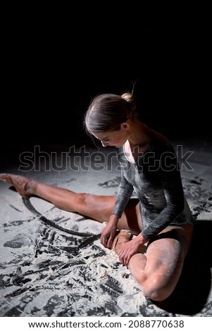 side view of exhausted young female ballet dancer in bodysuit sitting on floor with flour while resting after training in studio on black background, talented female ballerina sit relaxed.