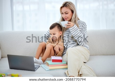 Smiling mom with cute lovely son using laptop watching cartoons. nice mum and preschool child sitting on sofa at home, watching video online, mom hugging son, drinking tea. in bright cozy room