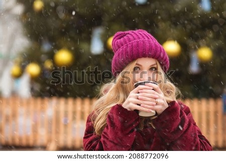 Charming blond woman wears red knitted cap drinking coffee at the winter fair. Empty space