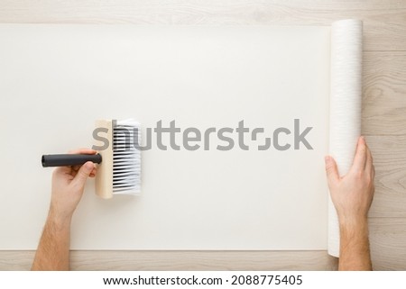 Young adult man hand using brush and applying glue on wallpaper sheet on wooden table background. Closeup. Repair work of home. Point of view shot.  Royalty-Free Stock Photo #2088775405
