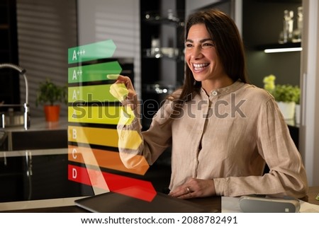 Young happy woman using futuristic innovative technology screen with augmented reality holograms to choose energy class efficiency rating consuming environment of electrical appliances at her home.