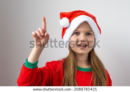 Portrait of little smart girl in Christmas red hat on white background. Child raised index finger up with great new idea. Inspiration motivation, New Year celebration, making wishes for Santa Claus.