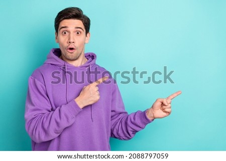 Photo of funny shocked young guy dressed violet sweatshirt pointing two fingers empty space isolated turquoise color background