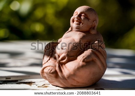 A laughing clay Buddha figurine in a sunny garden.