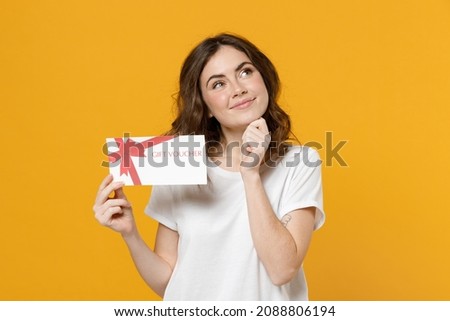 Young happy dreamful pensive wistful fun caucasian woman in white basic t-shirt point index finger on gift voucher flyer mock up prop up chin look aside isolated on yellow background studio portrait