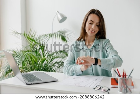 Young smiling happy successful employee business woman 20s in blue shirt look at smartwatch time sit work at workplace white desk with laptop pc computer at office indoors. Achievement career concept Royalty-Free Stock Photo #2088806242