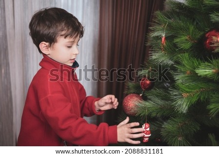 Close-up of a happy little boy in a red sports jacket decorates the Christmas tree with a big glass red ball and toy in the form of Santa Claus. Copy space. Home decoration for the holiday.