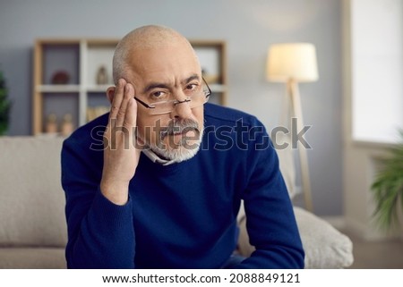 Portrait of strict reserved senior man in glasses sitting on couch with serious face, feeling skeptic, doubtful, uninspired and unexcited about bad idea or having secondhand embarrassment for somebody Royalty-Free Stock Photo #2088849121