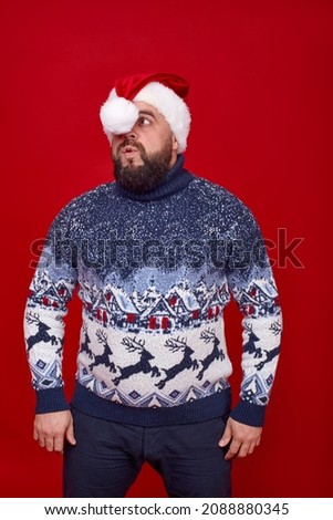 A funny man in a New Year's sweater is blowing on a New Year's cap on a red background.