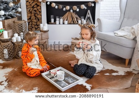 A boy and a girl in tiger cubs costumes are playing on the floor.Brother and sister in pajamas.The symbol of the new year is a tiger.Children in kigurumi pajamas drink cocoa and cookies