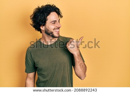Handsome hispanic man wearing casual green t shirt pointing thumb up to the side smiling happy with open mouth 