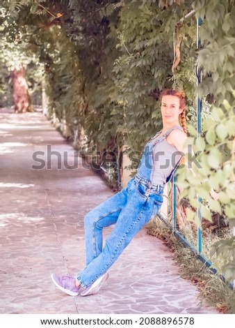 A lady in denim overalls is leaning on a metal fence in a green park alley on a summer day