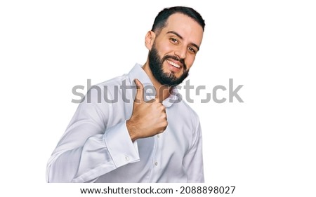 Young man with beard wearing business shirt doing happy thumbs up gesture with hand. approving expression looking at the camera showing success. 