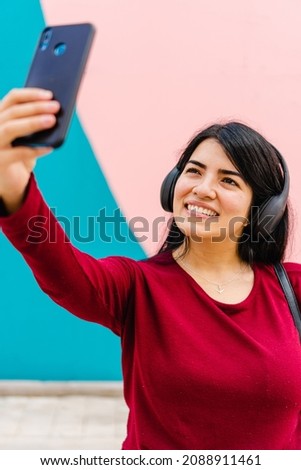 young latina female student listening to music or podcast with wireless headphones, while taking a selfie with her smart phone, colorful pink and blue background, vertical photo