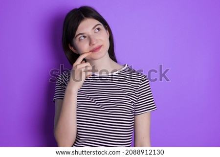 Lovely dreamy Caucasian woman wearing striped T-shirt isolated over studio background keeps finger near lips looks aside copy space.