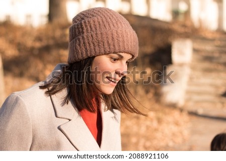 A girl with long dark hair in a coat and a knitted hat walks against the backdrop of an empty autumn park. Photo of a smiling model looking to the side.