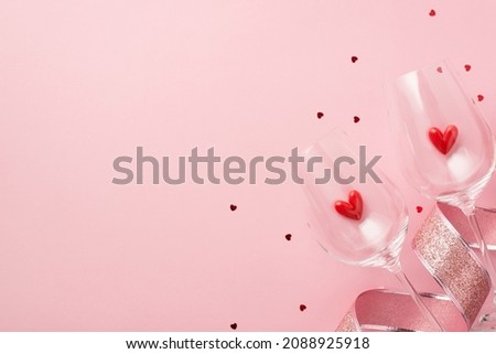 Top view photo of valentine's day decorations glitter pink curly ribbon red small hearts in two wineglasses and red heart shaped confetti on isolated pastel pink background with empty space