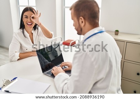 Young hispanic woman at the doctor smiling happy doing ok sign with hand on eye looking through fingers 