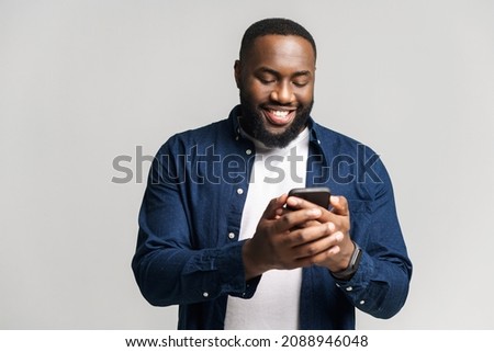 Optimistic African-American man wearing casual shirt texting on the smartphone, texting, smiling black guy enjoying using new mobile app isolated on grey