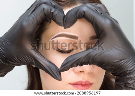 Permanent eyebrow makeup. Hands in black gloves make a heart, inside the eyes. Eyebrow care. Tattooing. Eyebrow coloring. Royalty-Free Stock Photo #2088956197
