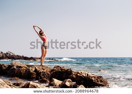 Happy young woman enjoying freedom with open hands on vacation while standing on the coast rocks enjoying sea and blue sky view