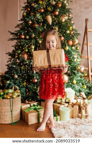 A little girl in a red dress with a gift box in her hands stands by the Christmas tree. Christmas Holidays. Christmas decor, atmosphere, winter background