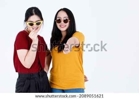 Portrait studio shot two young Asian cool fashionable stylish female friends model in casual outfit and fashion sunglasses stand smile look at camera show finger mini heart sign on white background.