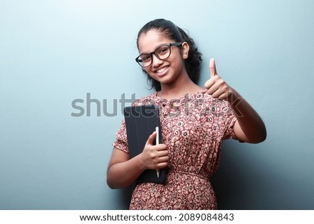 Portrait of a happy girl of Indian ethnicity with a tablet phone in hand shows thumbs up gesture  Royalty-Free Stock Photo #2089084843