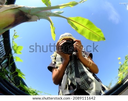 Amazing Vietnamese female photographer take photo by selfie via convex mirrors, woman sit on wooden chair, camera in hand, under banana tree and blue sky at home rooftop garden