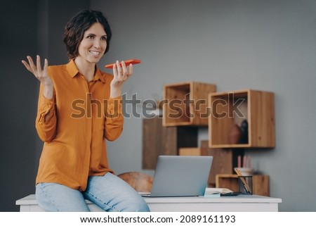 Happy joyful hispanic female using speakerphone or virtual voice assistant on mobile phone while working at home office, smiling emotional european woman recording audio message for colleague