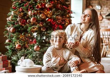 pregnant mom and little kid blond boy in beautiful knitted clothes sits in living room near Christmas tree in chalet with white small rabbits for family celebration of Christmas and New Year
