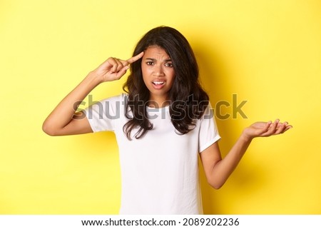 Portrait of angry and confused african-american girl, scolding someone for being stupid, pointing at head and shrugging, standing over yellow background