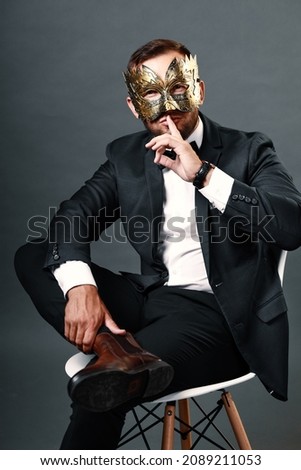 Man in business suit sits on chair wearing carnival mask on his face. Studio portrait on gray background. Young guy of 25-30 years hides his face under mask. Cunning and deceitful businessman Royalty-Free Stock Photo #2089211053