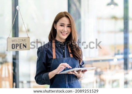 Smiling young Asian woman owner standing holding a tablet and open sign board. Looking at camera.