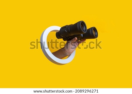 Woman's hand holding binoculars in a hole on a yellow background  Royalty-Free Stock Photo #2089296271