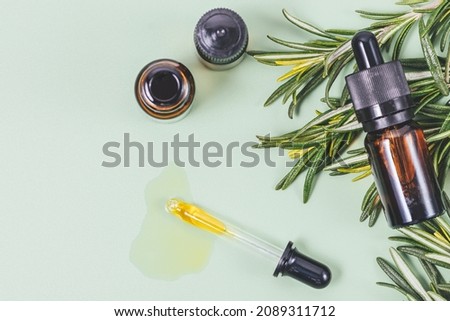 Rosemary essential oil flows from pipette next to bottles and fresh herbs of rosemary on green background, top view