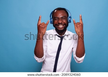 Smiling man full of energy doing rock and roll hand sign while listening to music in wireless headphones. Business man acting rebel doing hand sign while listening to loud music in earphones.