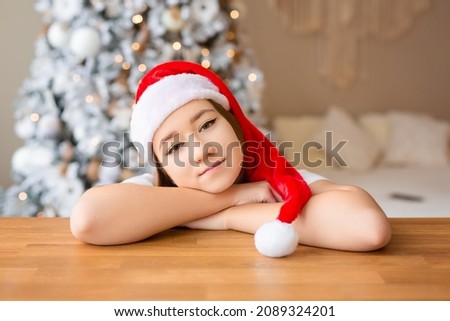 Girl in a red Santa hat looks out from behind the kitchen table, with a beautiful snow-covered Christmas tree in the background. High quality photo