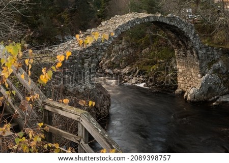 Long exposure of an old stone arch bridge over a flowing stream in Carrbridge, Cairngorms National Park, Highland of Scotland, United Kingdom, with yellow Autumn colour leaves on the foreground