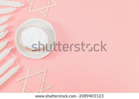 A donut with powder, white candles and wooden stars of David lie on the left against a pink background with copy space on the right, top view close-up. Hanukkah Celebration Concept.