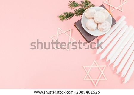 Powdered donuts in saucers, bible, nine white candles and three wooden stars of David lie on the right on a pink background with copy space on the left, top view close-up. Hanukkah Celebration Concept