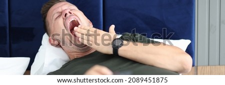 Young man lying in bed with remote control in his hand and yawning. Boring tv concept