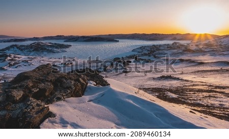 Winter dawn in Siberia. The sky is highlighted in orange. Glare from the sun's rays on the ice of a frozen lake. Campsite on the shore. In the foreground is a rock covered with sparkling snow. Baikal Royalty-Free Stock Photo #2089460134