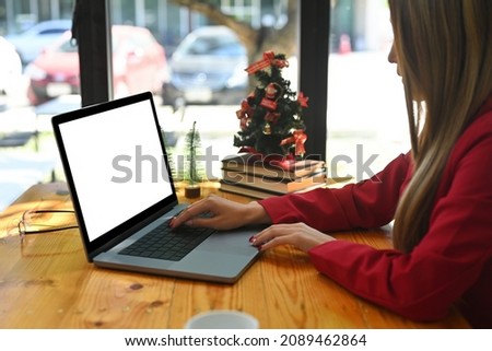 Photo of a young creative woman typing on a computer laptop at the wooden table surrounded by a coffee cup, eyeglasses, stack of books and Christmas tree.