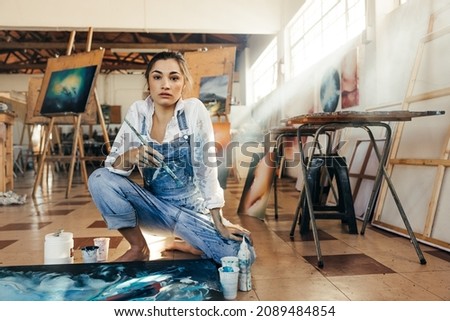 Inventive female painter looking at the camera in her art studio. Skilful artist making a blue painting for her new art project. Creative young woman working on the floor in her atelier. Royalty-Free Stock Photo #2089484854